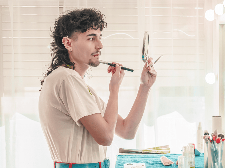 Young non-binary make-up artist applying makeup in front of a mirror with light bulbs in a bright room, focusing on diversity and inclusion in the beauty industry